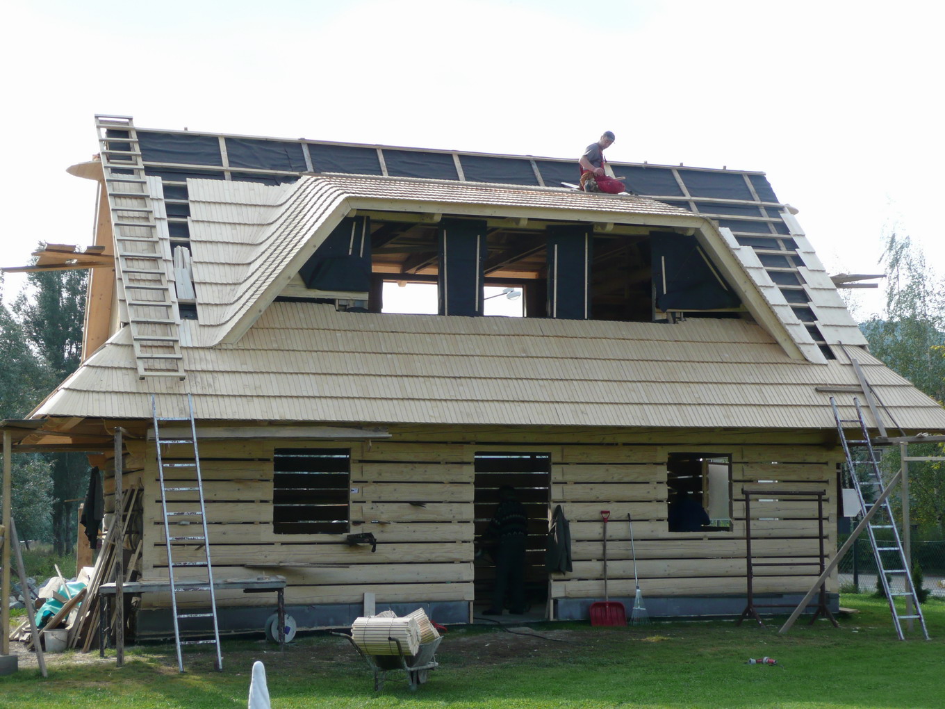 Budowa domu z drewnianych bali z charakterystycznym dachem pokrytym drewnianymi gontami; widoczny pracownik na dachu oraz drabiny opierające się o ściany.